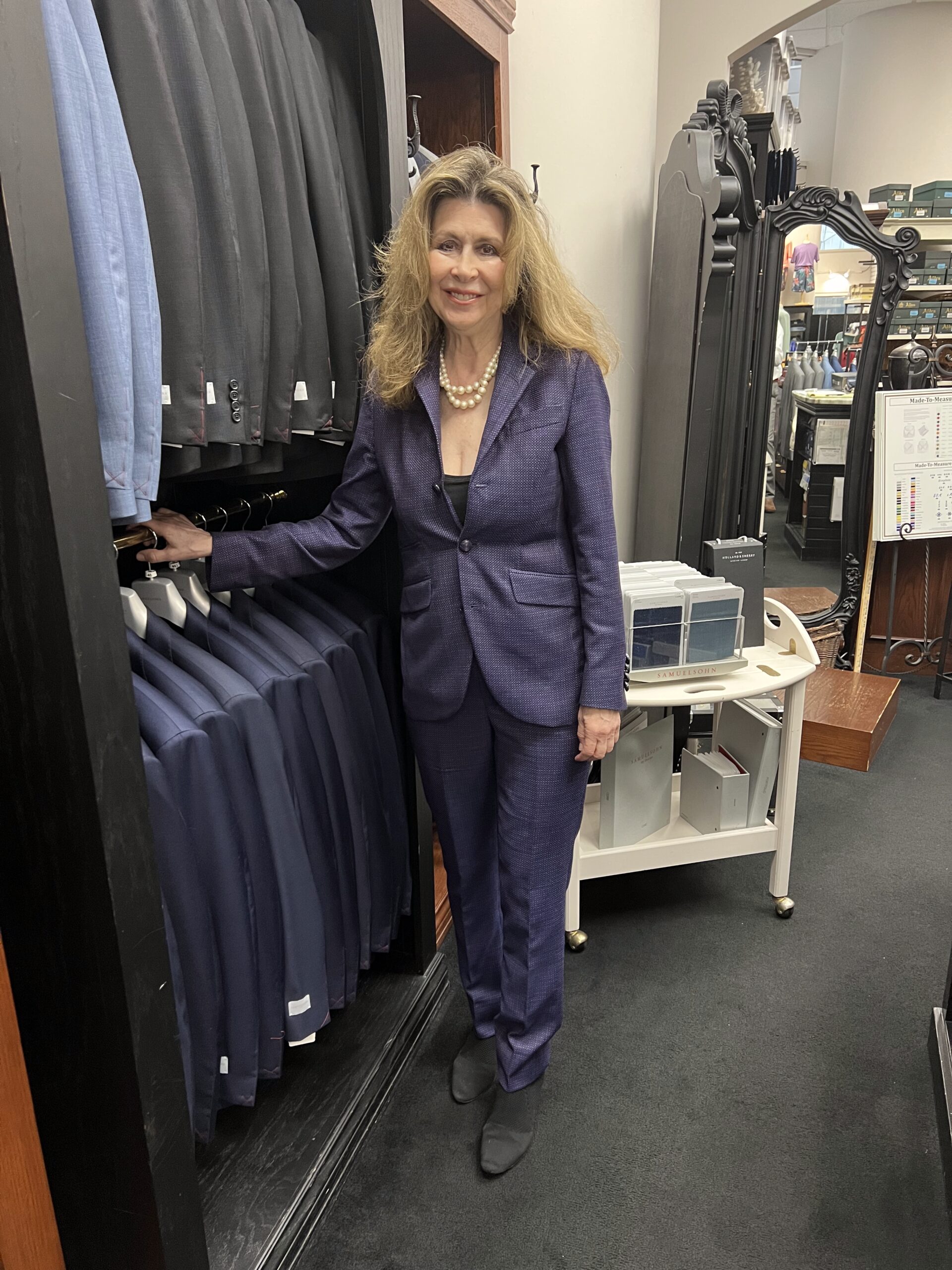 A woman in a tailored purple suit stands beside a rack of suits in a clothing store